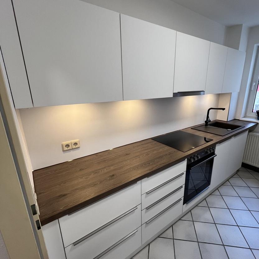 Einbauküche mit Holzarbeitsplatte von Tischlerei Krogull Schlichte Einbauküche mit weißen Fronten, einer eleganten Holzarbeitsplatte und integrierter Beleuchtung für eine moderne Küchenoptik.